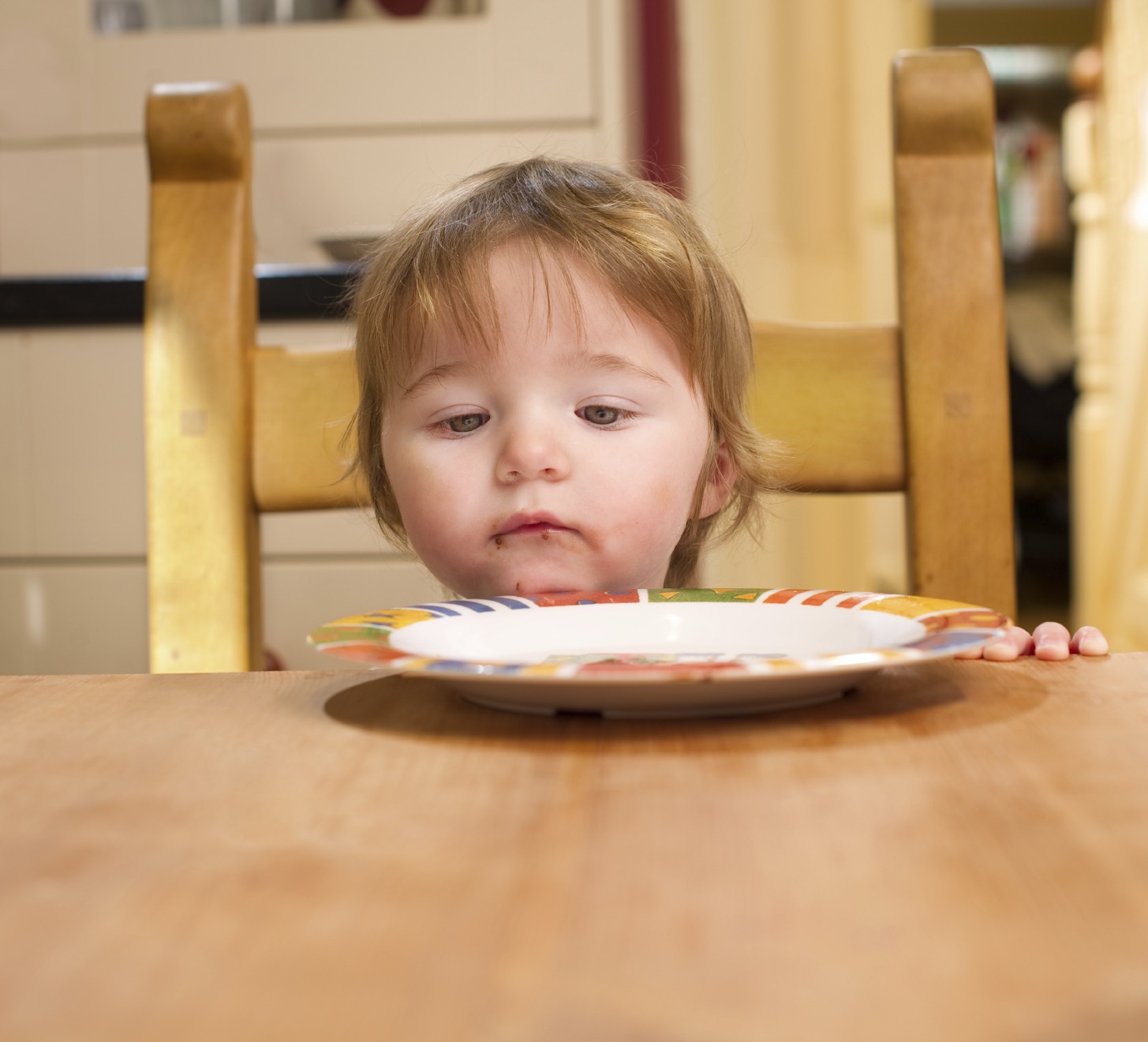 Young toddler sits in wood chair at wood table, looking at an empty plate