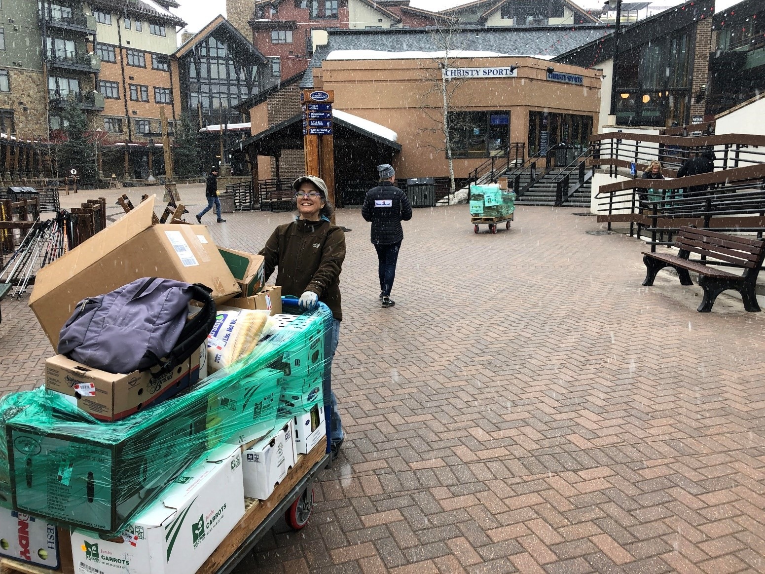Woman in brown jacket and tan baseball cap pushes large flat cart full of food boxes