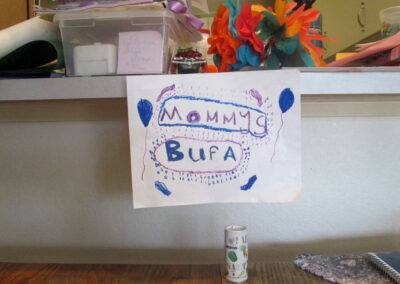 Sign in messy kitchen that says "Mommys bufa" (drawn by a child with crayons) underneath is a single salt shaker