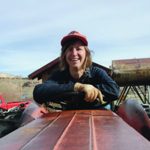 Image of woman smiling and leaning on a tractor. She's wearing working gloves and a red trucker hat