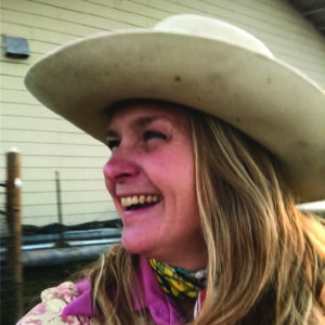 Headshot of Rachel Landis, she wears a white cowboy hat and a pink and white shirt. She's in profile, laughing.