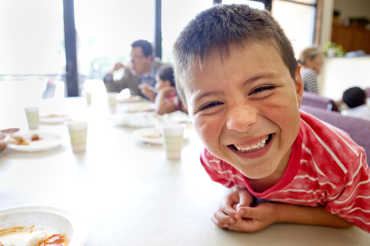 Young boy smiles at the camera while sitting at a table in a school cafeteria. Child nutrition is essential to strong foundations.